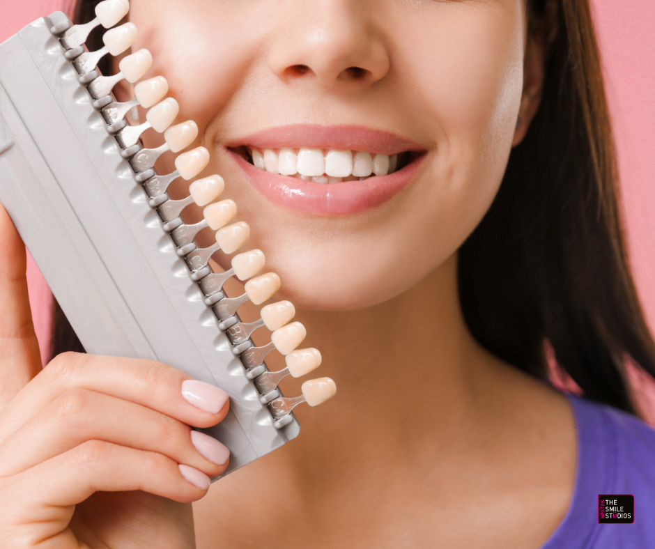 Close up of the bottom two-thirds of a young woman's teeth. She is holding up a set of false teeth. 